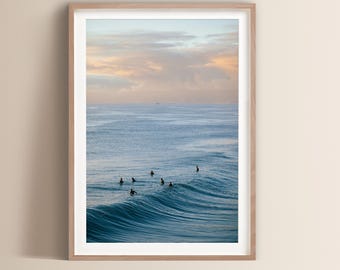 May include: A framed print featuring a serene ocean scene. The image captures a wave with several surfers in the water. The sky displays a soft blend of pastel colors, creating a tranquil and inviting atmosphere. The print is encased in a light-colored wooden frame.