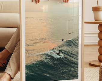 May include: A framed print featuring a serene ocean scene with surfers riding waves. The artwork captures the golden hues of a sunset, with the ocean waves in shades of teal and turquoise. The frame is a light wood color.