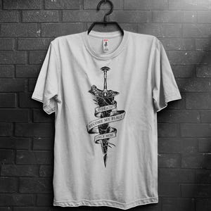 Può includere: T-shirt bianca con grafica nera di una spada e uno stendardo. Lo stendardo recita "O DEATH BECOME MY BLADE ONCE MORE". La maglietta è appesa a una gruccia nera contro un muro di mattoni scuri.