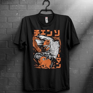 Könnte beinhalten: Schwarzes T-Shirt mit einem grafischen Design mit japanischem Text und einer Kettensägen-Charakter. Das Design enthält orange und weiße Elemente mit einer Cartoon-Illustration einer Kreatur. Das T-Shirt hängt an einem schwarzen Kleiderbügel.