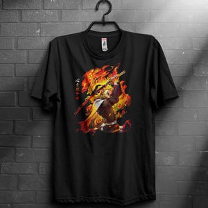 Puede incluir: Camiseta negra con una vibrante ilustración de un personaje envuelto en llamas, empuñando una espada. El diseño incluye texto japonés en naranja. La camiseta está colgada en una percha negra contra una pared de ladrillos oscuros.