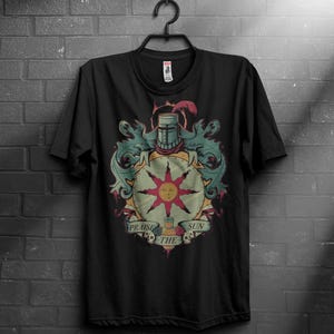 Può includere: T-shirt nera con un design colorato raffigurante un elmo da cavaliere, un emblema solare e la scritta "Praise the Sun". Il design include elementi turchese, gialli, rossi e beige. La maglietta è appesa a una gruccia nera contro un muro di mattoni.