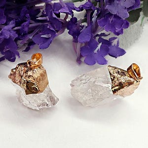 Puede incluir: Dos colgantes de cristal de cuarzo transparente con tapas y bucles de metal dorado. Los cristales son de corte rugoso e irregular. Flores moradas en el fondo, sobre una superficie blanca.