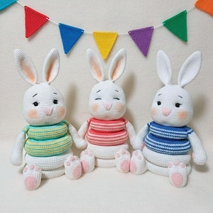 Può includere: Tre bambole di coniglietto bianche fatte a mano all'uncinetto con nasi e guance rosa, che indossano magliette a righe verdi, rosse e blu. I coniglietti sono seduti in fila, tenendosi per mano, con uno striscione colorato sullo sfondo.