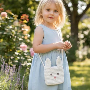 May include: A white crochet bunny-shaped purse with a long strap. The purse has two upright ears, two small brown eyes, and a pink nose. The girl is wearing a light blue dress.
