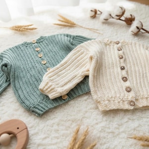 May include: Two knitted baby cardigans, one in a soft seafoam green and the other in creamy off-white. Both feature a ribbed texture, long sleeves, and wooden buttons. They are displayed on a fluffy white surface, creating a cosy, handmade feel.