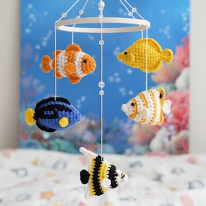 Könnte beinhalten: Handgefertigtes Babymobile mit sechs gehäkelten Fischen in verschiedenen Farben, darunter Orange, Gelb, Blau, Schwarz und Weiß. Die Fische sind an einem weißen, kreisförmigen Rahmen mit durchsichtigen Perlen aufgehängt und schaffen ein Unterwassermotiv.
