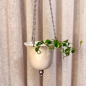 May include: A white ceramic hanging planter with a silver chain, holding a green plant with small leaves. The planter has a rounded bottom and a decorative silver ball. The background is a light-colored curtain.