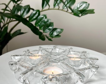 May include: A clear glass candle holder with a wavy, textured design. Three tea lights are placed in the holder, two of which are lit.