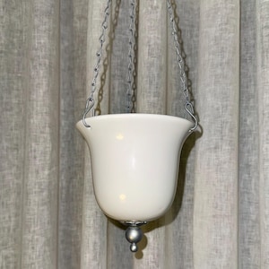 Può includere: Un lampadario bianco a forma di campana, sospeso da una catena d'argento. La lampada ha una base arrotondata con una piccola sfera decorativa d'argento sul fondo. Lo sfondo è una tenda chiara e strutturata.