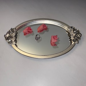 Puede incluir: Una bandeja ovalada con espejo de tono plateado que presenta motivos decorativos de rosas de metal en ambas asas. Tres pétalos de flores rosas secos y un anillo plateado con una piedra central se sitúan sobre la superficie reflectante.