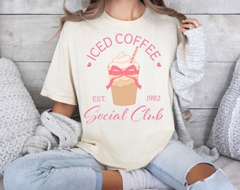 May include: A woman wearing a light beige t-shirt with a pink iced coffee graphic and the text "ICED COFFEE EST. 1982 Social Club".