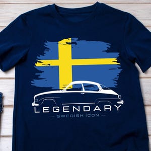 May include: Navy blue t-shirt featuring a white outline of a classic car with the text "LEGENDARY - SWEDISH ICON" below a stylized Swedish flag. The shirt is displayed on a wooden surface with jeans, sunglasses, and sneakers.