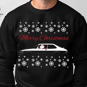 May include: Black sweatshirt with a white and black car graphic, snowflakes, and the text "Merry Christmas" in red. The car has Santa Claus in the driver's seat. A festive holiday-themed design.