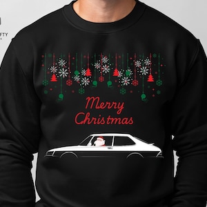 May include: Black sweatshirt with a Christmas design. The design includes red and green ornaments, snowflakes, and the words "Merry Christmas." A white and black car with Santa Claus in the driver's seat is featured below the text.