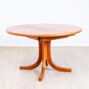 Könnte beinhalten: Ein runder Esstisch aus Holz mit einer glatten, hellbraunen Oberfläche. Der Tisch verfügt über einen zentralen Säulenfuß, der sich in vier geschwungene Beine verzweigt und eine stabile Basis für die runde Tischplatte bietet.