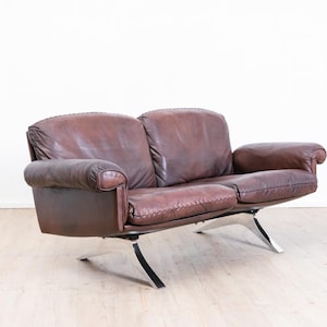 Peut inclure: Un canapé deux places recouvert de cuir marron foncé avec des coutures apparentes le long des coussins et des accoudoirs. Le canapé présente un design bas soutenu par quatre pieds incurvés en métal chromé poli qui s'étendent vers l'extérieur.