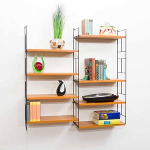 Può includere: Una libreria a parete in stile moderno di metà secolo con quattro ripiani in legno e una struttura in metallo nero. I ripiani espongono libri, vasi decorativi e una pianta in un vaso a fantasia. L'unità è contro un muro bianco.