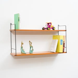 Può includere: Un ripiano a muro in legno con supporti in metallo nero. Il ripiano ha due livelli ed è decorato con libri, un'auto giocattolo e due piccole sculture.
