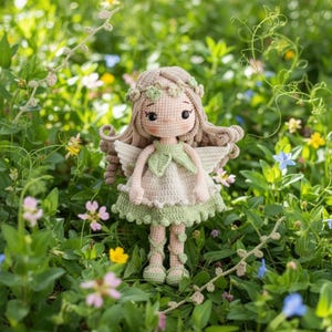 Può includere: Una bambola all'uncinetto fatta a mano in un giardino. La bambola ha lunghi capelli castani, una corona floreale e un vestito verde e crema. Ha ali delicate ed è circondata da verde e fiori.