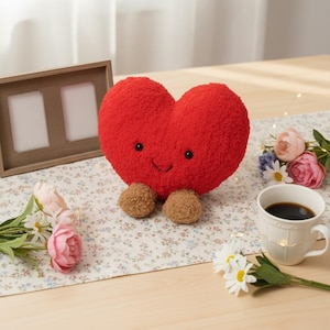 Peut inclure: Un coussin en forme de cœur rouge avec un visage souriant et de petits pieds bruns. Le coussin est posé sur une nappe à motifs floraux, à côté d'un cadre photo en bois, de fleurs et d'une tasse de café.