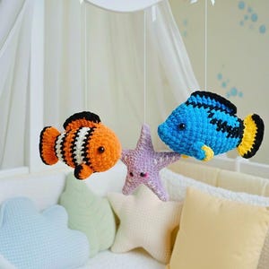 May include: A handmade baby mobile featuring three crocheted sea creatures: an orange and white clownfish, a blue fish with black stripes, and a lavender starfish. The mobile hangs above a crib with pastel-colored pillows.
