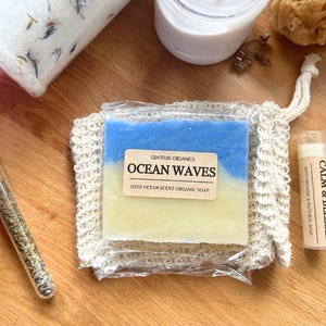 以下が含まれることがあります： 木製の表面に置かれたスパ製品の詰め合わせ。 「Ocean Waves」の石鹸、 「Calm & Blessed」と書かれたリップバーム、ガラスバイアル、バスソルトの瓶が含まれています。 石鹸は青と黄色で、リップバームはベージュ色のチューブに入っています。