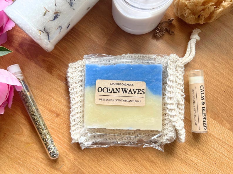 以下が含まれることがあります： 木製の表面に置かれたスパ製品の詰め合わせ。 「Ocean Waves」の石鹸、 「Calm & Blessed」と書かれたリップバーム、ガラスバイアル、バスソルトの瓶が含まれています。 石鹸は青と黄色で、リップバームはベージュ色のチューブに入っています。
