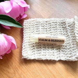 以下が含まれることがあります： 「CALM & BLESSED」の文字が入ったリップバームチューブが、ベージュ色のニットポーチの上に置かれています。背景にはピンク色のシャクヤクと緑の葉があります。リップバームは「GRATITUDE ORGANICS」製です。背景は木製の表面です。