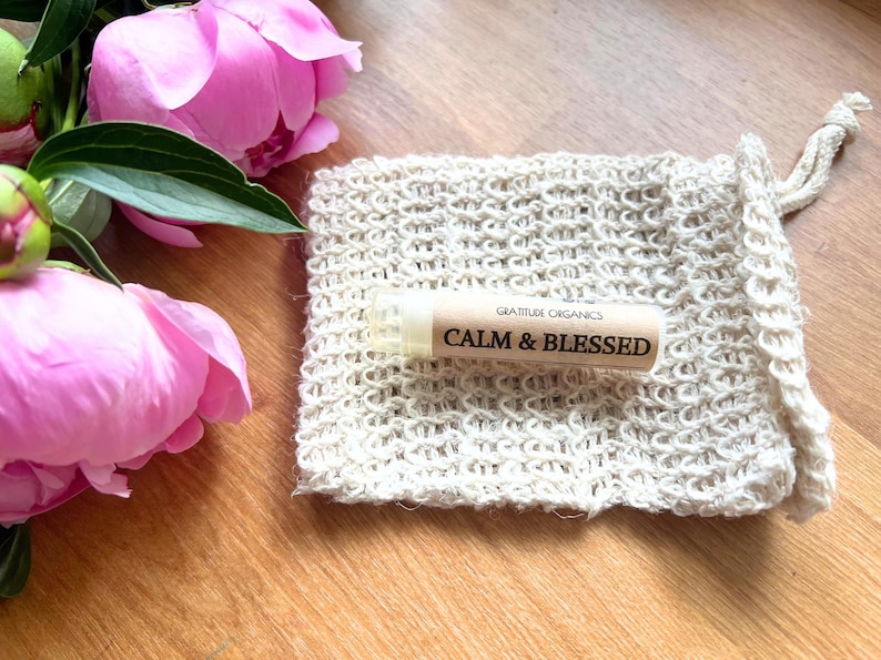 以下が含まれることがあります： 「CALM & BLESSED」の文字が入ったリップバームチューブが、ベージュ色のニットポーチの上に置かれています。背景にはピンク色のシャクヤクと緑の葉があります。リップバームは「GRATITUDE ORGANICS」製です。背景は木製の表面です。