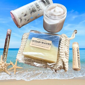 以下が含まれることがあります： 「Ocean Waves」の石鹸、クリームの瓶、バスソルトのチューブ、「Calm & Blessed」と書かれたリップバームなど、バス＆ボディ製品のコレクション。ヒトデのあるビーチに配置されています。