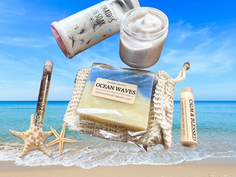 以下が含まれることがあります： 「Ocean Waves」の石鹸、クリームの瓶、バスソルトのチューブ、「Calm & Blessed」と書かれたリップバームなど、バス＆ボディ製品のコレクション。ヒトデのあるビーチに配置されています。