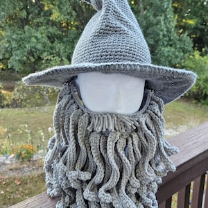May include: A gray crocheted wizard hat with a pointed top and wide brim, paired with a matching crocheted beard. The hat and beard are displayed on a white mannequin head, set against a blurred outdoor background.