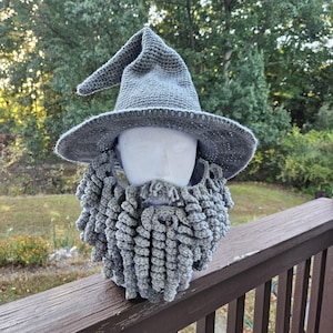 May include: A gray crocheted wizard hat with a pointed top and wide brim, paired with a matching crocheted beard. The beard has a textured, looped design. The hat and beard are displayed on a white mannequin head.