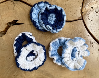 May include: Three crocheted hair scrunchies with ruffled edges. One scrunchie features dark navy blue and light blue yarn. Another is white with a dark navy blue border. The third is white with a light blue border, all with a circular shape.
