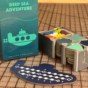May include: A Deep Sea Adventure board game box in teal with a submarine graphic, a silver 3D-printed organizer containing stacks of hexagonal tiles in various shades of blue and green, and a dark blue submarine-shaped score tracker board.