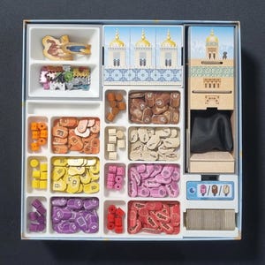 May include: A white plastic organizer with multiple compartments filled with colorful game pieces. The organizer has a blue and white design with a picture of a building on the front. The game pieces include small wooden pieces, plastic tiles, and dice.