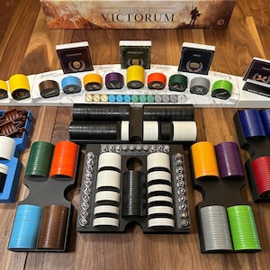 May include: A board game set with various components, including colorful chips, dice, and card holders. The game pieces are arranged on a wooden surface, with a box labeled "Hoplomachus Victorum" in the background.