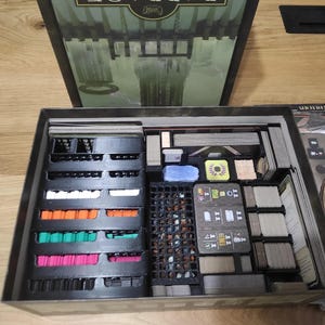 May include: A black and gray board game organizer with multiple compartments for storing game pieces and cards. The organizer is filled with various colored pieces and cards.