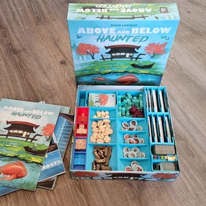 May include: A board game box set titled "Above and Below Haunted" with a light blue exterior. The box is open, revealing game components in a blue tray. Rule books with the same title and artwork are visible.