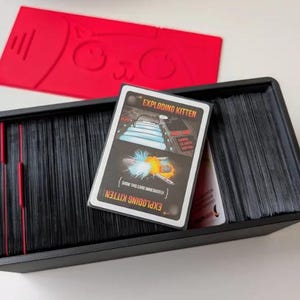 May include: A black rectangular card storage box filled with stacks of black-edged playing cards. An Exploding Kitten game card is placed on top of the stacks. A separate red plastic lid featuring a stylized cat face design is positioned above the box.