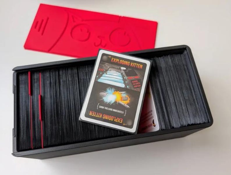 May include: A black rectangular card storage box filled with stacks of black-edged playing cards. An Exploding Kitten game card is placed on top of the stacks. A separate red plastic lid featuring a stylized cat face design is positioned above the box.