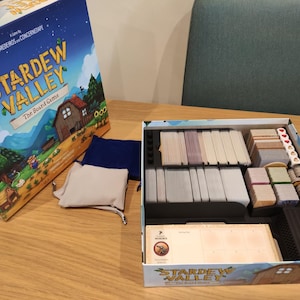 May include: An open box of the board game "Stardew Valley" shows organised game components. The box art depicts a rural scene with the game's title. Inside are card stacks, tokens, dice, and drawstring bags, indicating a strategy game.