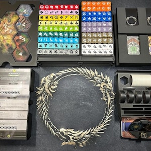 May include: A board game organizer with various components. Includes dice, game tiles, cards, and tokens. The set features a large, ornate circular emblem and several black storage trays. The color palette includes black, white, and various colors for the dice.