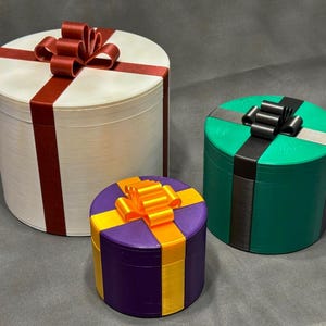 May include: Three cylindrical gift boxes of varying sizes. The largest is white with a red ribbon and bow. The medium box is green with a black ribbon and bow. The smallest is purple with a yellow and orange ribbon and bow.