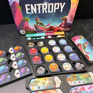 May include: The board game Entropy features a box with colorful alien landscapes and creatures. The set includes black plastic trays containing various circular tokens with planetary designs, hexagonal tiles with space imagery, and small resource cubes.