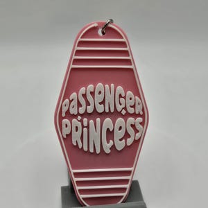 Puede incluir: Llavero de motel rosa con texto blanco que dice "Passenger Princess".