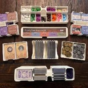 May include: A collection of white plastic game organizers filled with various game components, including cards, tokens, and dice. The organizers are arranged on a dark wooden surface, showcasing a variety of colors and shapes.