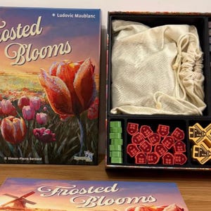 May include: The board game "Frosted Blooms" is displayed, featuring a box with a tulip illustration and the game title. Inside the box are game pieces, cards, and a drawstring bag. The game's theme appears to be related to flowers and windmills.