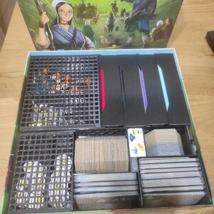 May include: A black and green game box with a foam insert for organizing game pieces. The box lid features a colorful illustration of a group of people in a field. The text on the lid reads "OSADNICY Z KRWI I KOSCI".